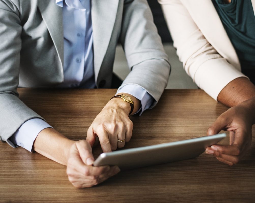 Two formally dressed individuals each hold on side of an electronic tablet, with one pointing something out on the screen to the other.